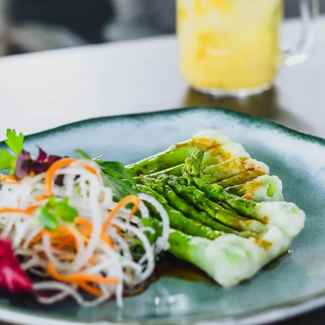 Teller mit grünem Spargel und Gemüsestreifen, dahinter eine hausgemachte Limonade im Henkelglas